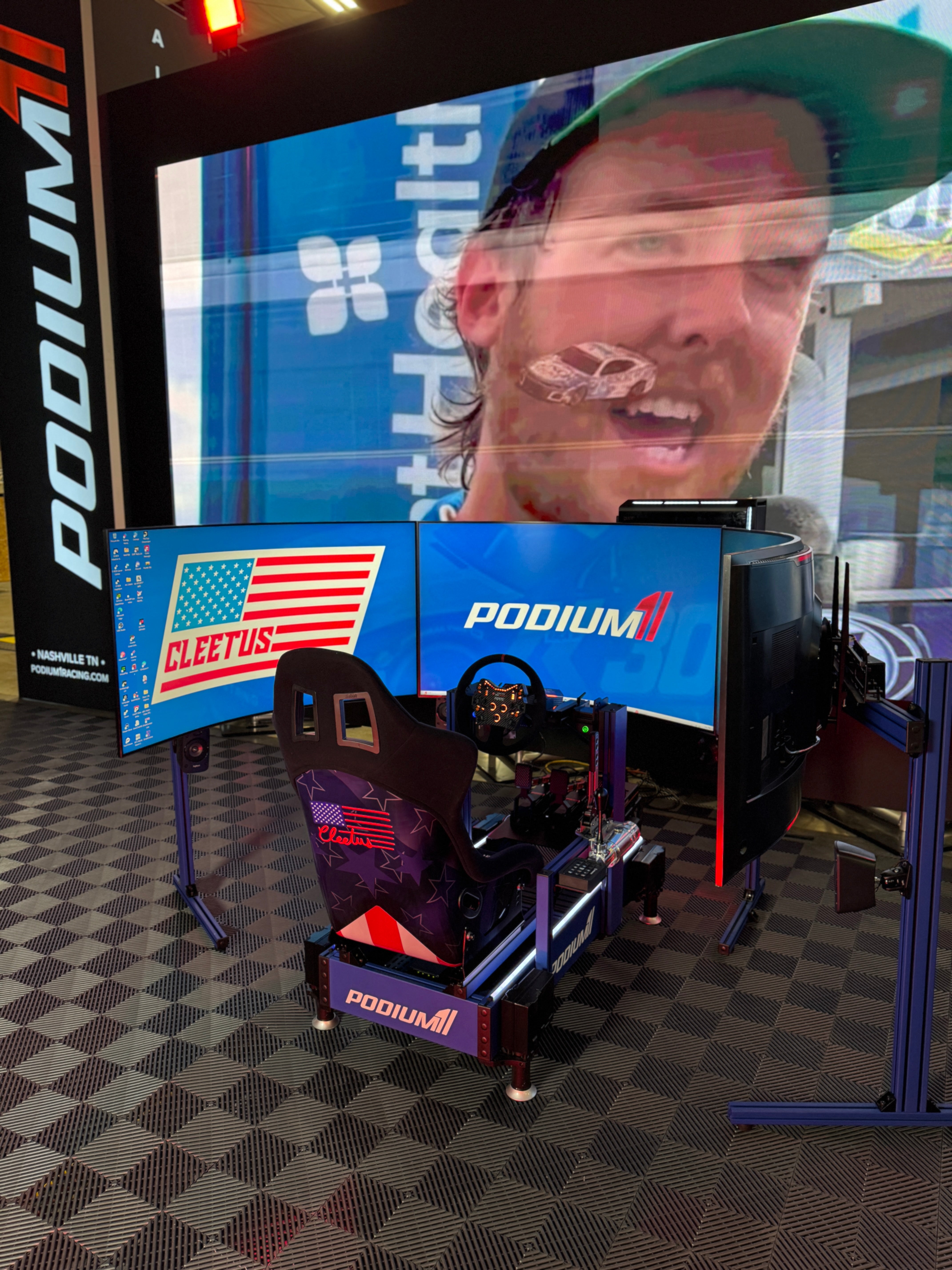 Racing simulator setup with 'Podium' branding and a person wearing a cap in front of a screen.